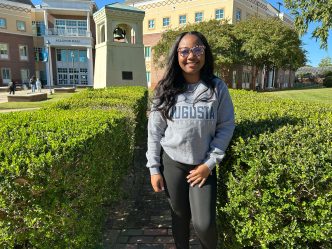 A young woman in glasses and an Augusta University sweater smiles at the camera on AU's campus.