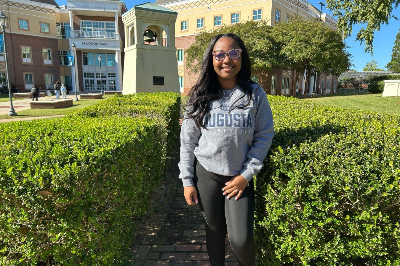 A young woman in glasses and an Augusta University sweater smiles at the camera on AU's campus.