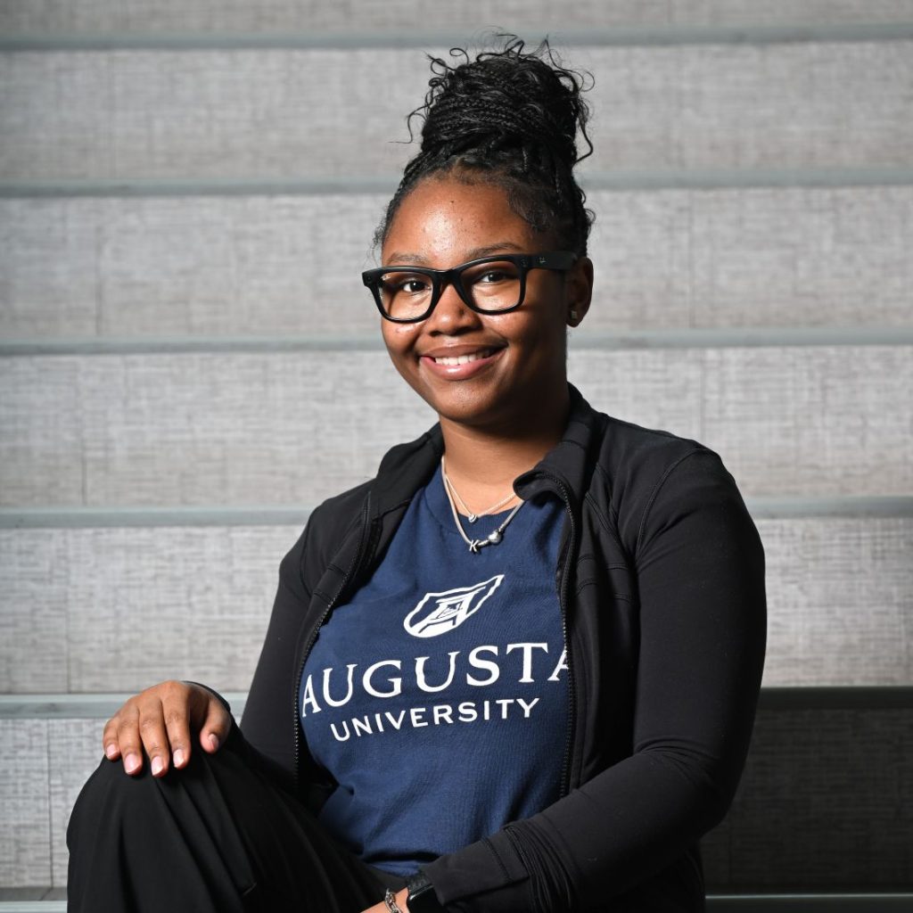 A young woman in glasses and an Augusta University shirt smiles at the camera