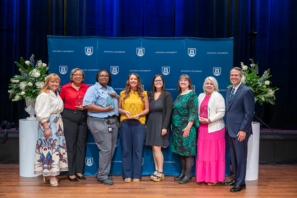 A group of women and one man stand together posing for a photo, some of the women holding glass awards.