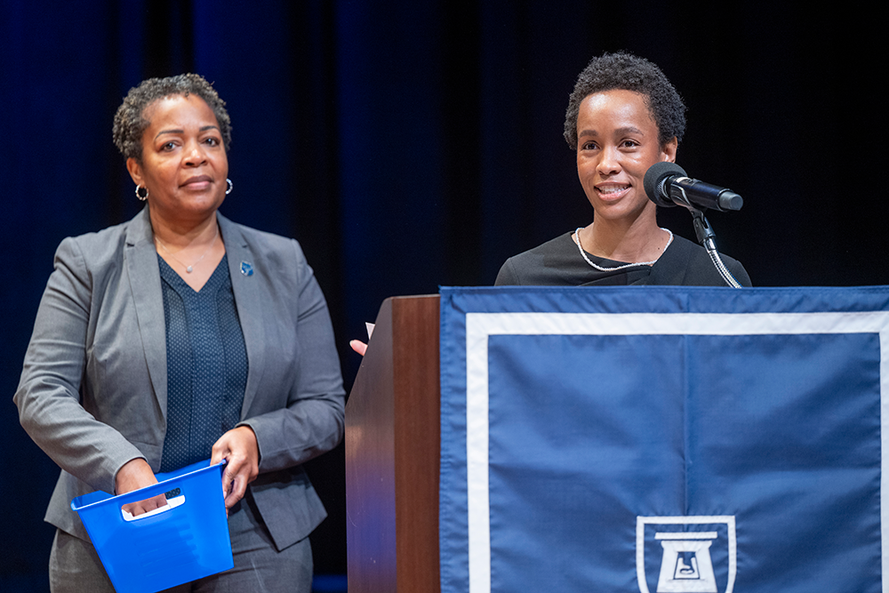 Two women stand behind a podium while one carries a blue box.