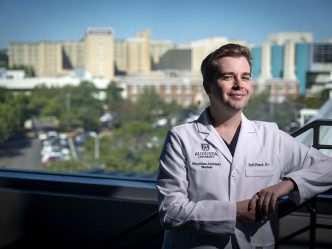 A man standing in a white coat with buildings in the landscape behind him
