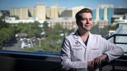 A man standing in a white coat with buildings in the landscape behind him