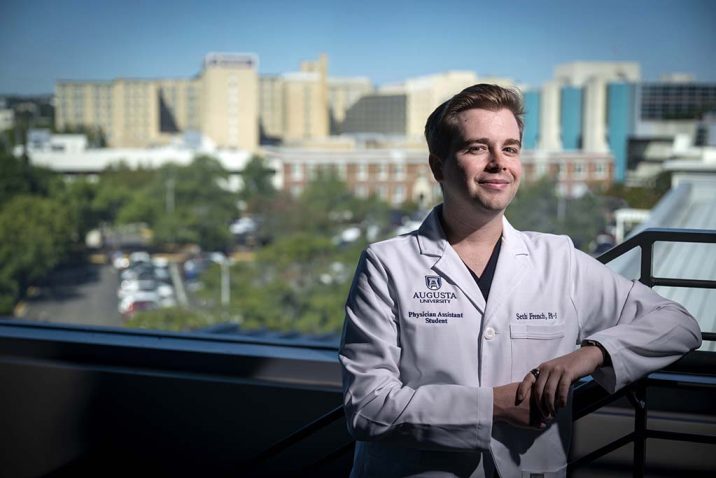 A man standing in a white coat with buildings in the landscape behind him