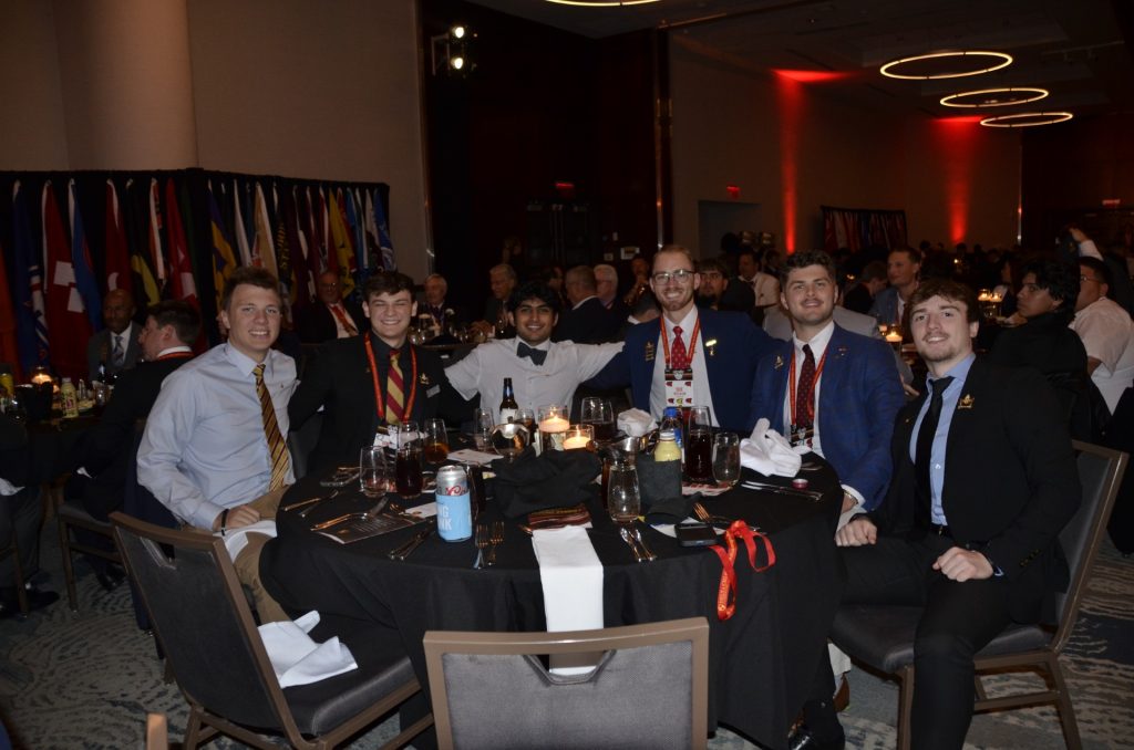 Several college-aged men gathered around a table smile for a photo.