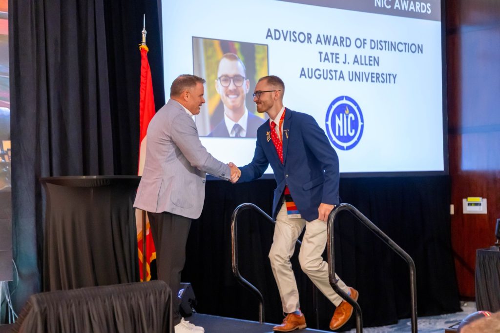 A college-aged man accepts an award while shaking another man's hand.