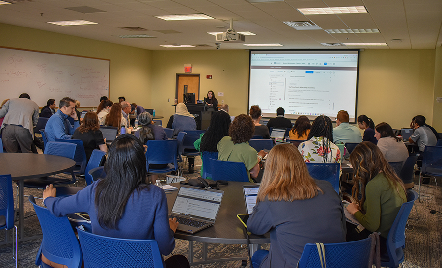 woman sitting at a podium speaking to people sitting at tables working on their laptops. 