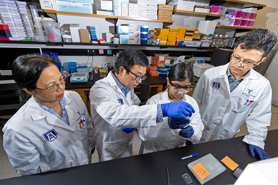 A group of four scientists gather around a lab bench to work on an experiment. Two of the scientists are working with tools, while the other two are watching.